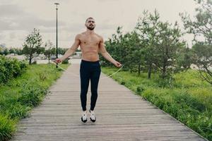 Sporty muscular man uses skipping rope for cardio training outdoor, has perfect body, breathes fresh air and demonstrates endurance, works out self development. Athlete guy with sport equipment photo