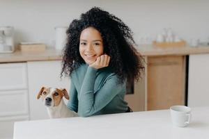 Photo of pleasant looking woman dressed in casual wear, has curly bushy hair, poses against kitchen interior with pedigree dog, has morning coffee, enjoys weekend, smiles pleasantly at camera