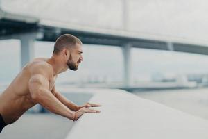 Sideways shot of handsome muscular man with thick beard does push ups exercise has workout outdoor leans on bridge fence engaged in physical activity poses with bare torso has strong muscles photo