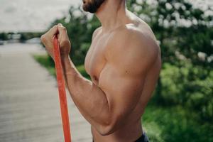 Cropped shot of unrecognizable muscular man has workout biceps exercise stands in profile shows strong muscles, uses resistance band, strives for strong body. Bodybuilding, motivation concept photo