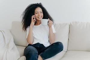 Cheerful Afro American woman has coffee break in living room, sits on couch, calls friend via modern gadget, has happy smile, thoughtful expression, discusses something pleasant. Technology, lifestyle photo