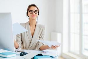 Busy successful female manager works with papers in office, poses at desktop, wears spectacles and formal outfit, busy preparing report, looks confidently at camera. Businesswoman reviews documents photo