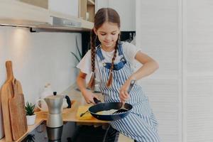 Little girl with pigtails wears apron, learns to cook, poses near stove, prepares fried eggs for breakfast, helps parents with cooking, busy at modern kitchen. Children, culinary, food concept photo