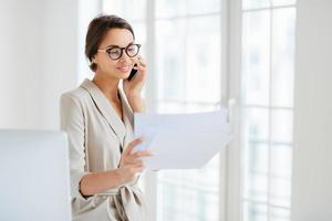 Positive woman in formal clothes, calls to business partner, discusses financial startup project, reads information from papers, wears optical glasses, stands in office, works at international company photo