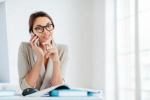 Photo of female employee has cheerful expression, talks via smartphone, hold pen, sits at desktop with papers and notepads, computer, poses against office interior, wears spectacles, elegant clothes