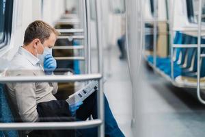 Prohibition of free movement. Man in formal clothes, coughs and has symptoms of coronavirus, wears protective medical mask and gloves, reads newspaper, poses in subway or metro, commutes to work photo