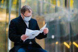 Coronavirus epidemic in city. Horizontal shot of serious man reads newspaper attentively, poses against blurred building background, wears mask for spreading of coronavirus. Businessman with press photo