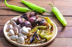 Grilled garlic green spicy pepper and red onion putting in wooden plate on wooden floor prepared for chili paste cooking in Thai style. photo