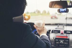 el hombre se apresura a conducir un automóvil usando el móvil y come comida rápida peligrosamente foto