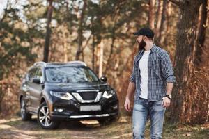 chico maduro dar un paseo. hombre barbudo cerca de su nuevo auto negro en el bosque. concepto de vacaciones foto