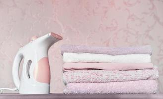 portable electric garment steamer and pile of folded towels on ironing board. housekeeping and householding, chores concept. pink and lilac clothes. photo