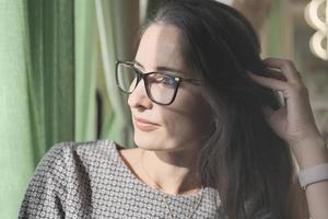 asian or middle east woman wearing eyeglasses looking aside and touching her hair. getting rest or break in office, enjoying sunshine. beautiful thoughtful calm woman. photo