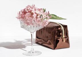 high crystal glass for champaign with flowers in it and shining glossy female hand bag or purse on white background. holiday party, selebration of birthday concept. photo