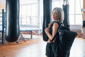 rubia deportiva lista para el entrenamiento. hembra adulta con bolsa negra y auriculares en el gimnasio foto