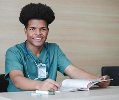 Portrait of medical student sitting at desk looking at camera smiling. Young African American doctor read book, and documents preparing for exams. Professional health care and medicine occupation. photo