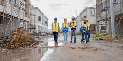 Team of engineers and workers wear safety clothing are walking through wet street in residential housing construction site. Engineering teamwork talking, discussing building project development.