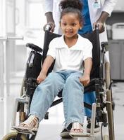 Portrait of cute mixed race kid sitting on wheelchair in hospital room with her doctor smiling. Little girl with physical disability is sick and come to clinic for professional medical treatment