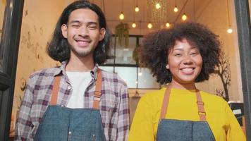Zwei junge Startup-Barista-Partner mit Schürzen stehen an der Tür eines ungezwungenen Cafés, die Arme verschränkt, schauen mit einem einladenden Lächeln in die Kamera, glücklich und fröhlich mit Jobs im Café-Service, KMU-Unternehmer. video