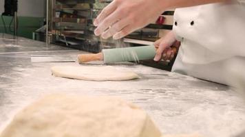 câmera lenta e close-up, chef em uniformes de cozinheiro branco com aventais polvilhe farinha, amassar massa de pastelaria com rolo, preparar pão, tortas e comida de padaria fresca na cozinha de aço inoxidável do restaurante. video