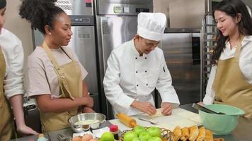 keukencursus, senior mannelijke chef-kok in kookuniform leert jonge kooklesstudenten om deegdeeg te kneden en te rollen, ingrediënten voor bakkerijproducten te bereiden, fruittaarten in roestvrijstalen keuken. video