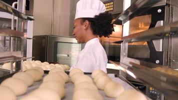 chef afroamericana profesional con uniforme de cocinera blanca, guantes y delantal haciendo pan con masa de pastelería, preparando comida fresca de panadería, horneando en el horno en la cocina de acero inoxidable del restaurante. video