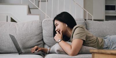Asian woman lying on the sofa in the living room listening to music on a laptop device woman relaxing on the sofa at home View messages on the computer screen on the device. communication concept photo