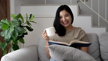 Young relaxed and happy asian woman in casual outfit reading book and drinking tea while relaxing on sofa in cozy living room at home photo