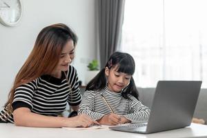 Niña pequeña asiática aprendiendo clases en línea en casa con su madre. el niño en edad preescolar usa una computadora portátil para hacer la tarea, la escuela en casa del maestro de la escuela por internet remoto digital con el apoyo de mamá. foto