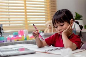 Little Asian girl sitting alone and looking out with a bored face, Preschool child laying head down on the table with sad bored with homework, spoiled child photo