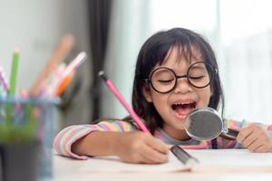 Student little Asian girl writing with a magnifying glass looking at the camera. sweet kids sitting in the living room at home. family activity concept. photo