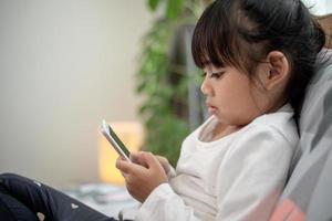 linda niñita viendo videos en un smartphone con una cara sonriente sola en la cama, un niño usando un teléfono móvil con una cara feliz en casa. quédese en casa cuarentena coronavirus covid-19 prevención de pandemias. foto