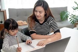 Asian little young girl kid learning online class at home with mother. Preschool child use laptop computer do homework, homeschool from school teacher by digital remote internet with support from mom. photo