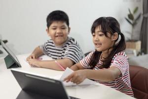 los niños y niñas asiáticos disfrutan del aprendizaje en línea tomando notas y usando tabletas en casa. foto