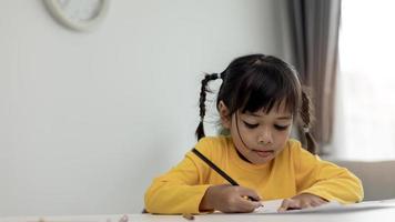 niño asiático estudiante de regreso a la escuela o niña sonrisa escribir una nota o dibujar con lápiz y leer en una tableta de computadora haciendo la tarea para pensar en una nueva idea o que la gente aprenda de casa foto