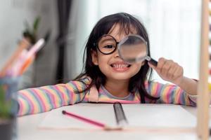 Student little Asian girl writing with a magnifying glass looking at the camera. sweet kids sitting in the living room at home. family activity concept. photo