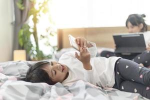 Cute little kid girl watching video on smartphone with smiley face alone on the bed, child using mobile phone with happy face at home. Stay at home quarantine coronavirus COVID-19 pandemic prevention. photo