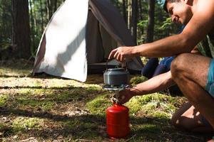 Cooking, heating a tourist kettle on a portable gas burner with a red gas cylinder. Camping, a man cooks breakfast outdoors. Summer outdoor activities photo