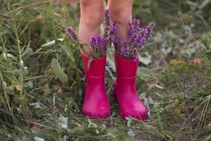 botas de goma rojas a los pies de una niña con un ramo de flores silvestres. flores en una bota, horario de verano. verano, libertad, naturaleza, campo, hierba verde en el campo foto