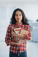 Vertical photo of attractive young woman standing in the office and holding books and documents