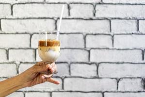 Coffee Macchiato and milk layer with ice in champagne glass was held by Asian grandmother hand in front of white brick wall wallpaper. photo