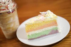 Slided rainbow cake decorate with unicorn horn on top next to coffee frappe. The cake is in a white disk on the wooden table and blur background. photo