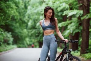 Caucasian ethnicity. Female cyclist standing with bike on asphalt road in the forest at daytime photo