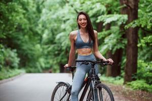 Beautiful abs. Female cyclist standing with bike on asphalt road in the forest at daytime photo