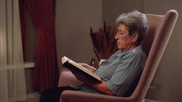 velha lendo um livro. mulher lendo um romance em casa à noite usando seus óculos. ele fecha o livro e olha pela janela. video