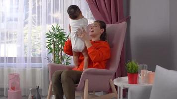 madre con su bebé. la madre sentada frente a la ventana cuida a su bebé en sus brazos y juega. Este es un video en cámara lenta.