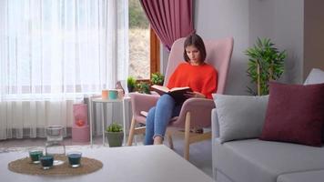 Frau, die ein Buch liest. Frau in orangefarbener Kleidung, die zu Hause ein Buch in einem rosafarbenen Sessel liest. Frau, die das neu gekaufte Buch im Wohnzimmer des Hauses untersucht. video
