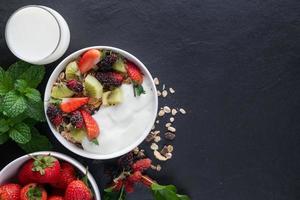 bowl of oat granola with yogurt, fresh mulberry, strawberries, kiwi mint and nuts on the black rock board for healthy breakfast, top view, copy space, flat lay. Healthy breakfast menu concept. photo
