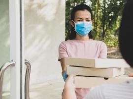 delivery service in t-shirt, in protective mask and gloves giving food order, holding three pizza boxes in front house, woman accepting delivery of boxes from delivery man during COVID-19 outbreak. photo