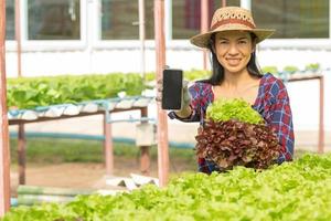 Asian woman farmers working using mobile in vegetables hydroponic farm with happiness. Portrait of woman farmer checking quality of green salad vegetable with smile in the green house farm. photo