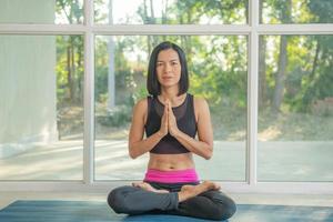 Young attractive woman practicing yoga, sitting in Padmasana, exercise, Lotus pose, namaste, working out, wearing sportswear,  pants black, indoor full length, near floor window. photo